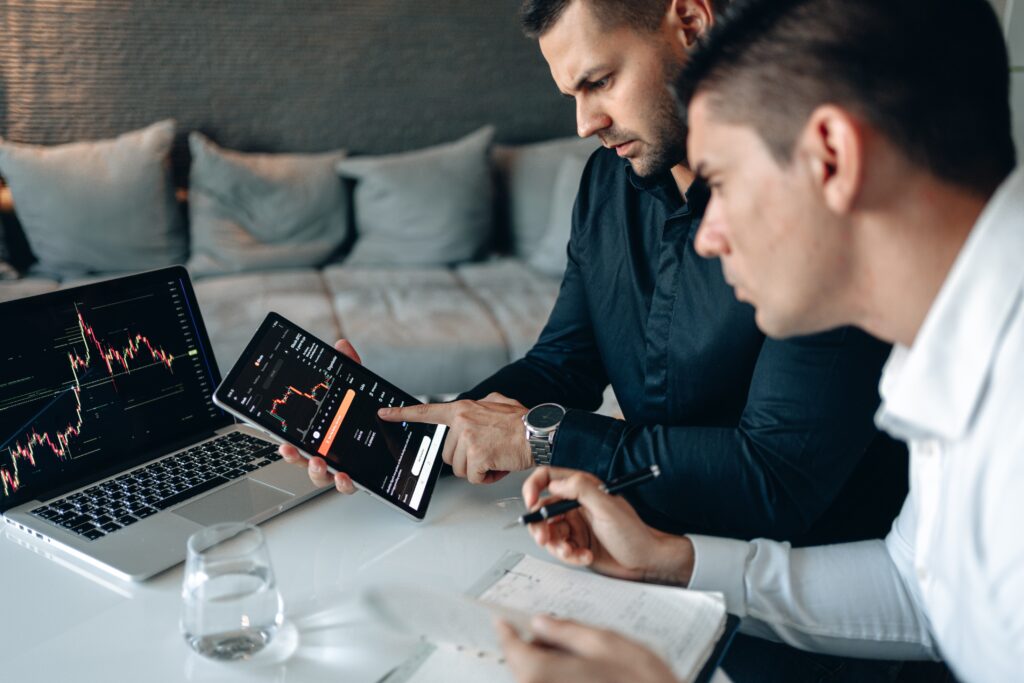two men looking over a site’s analytics on a laptop, one holding an iPad and another holding a pen and a notebook