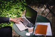 the hand of a man hovering above his laptop placed beside a cup of coffee, airpods, pens, a notebook, and documents on a small table outdoors