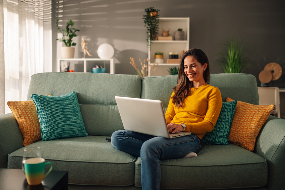 Smiling caucasian woman using laptop while sitting on a mint couch at home. Happy female shopping or chatting online in social network, typing blog, freelancer working on a project